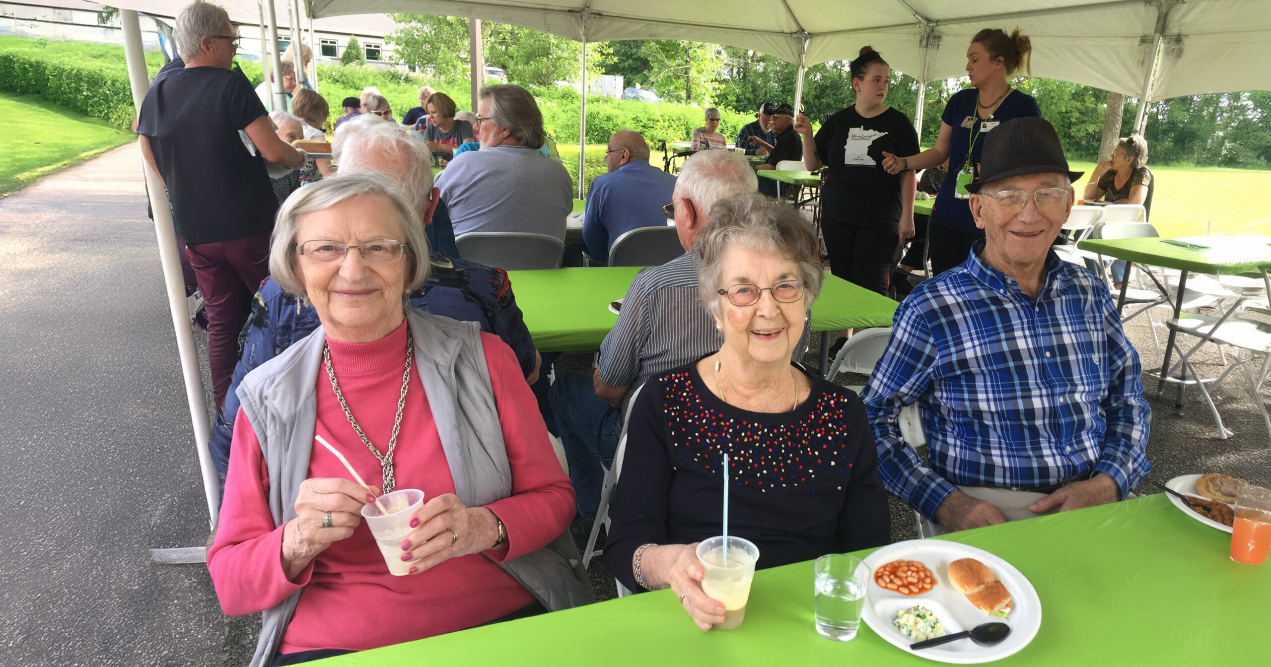 3 senior adults at an outdoor celebration with food sitting at folding tables on a sunny day under a tent
